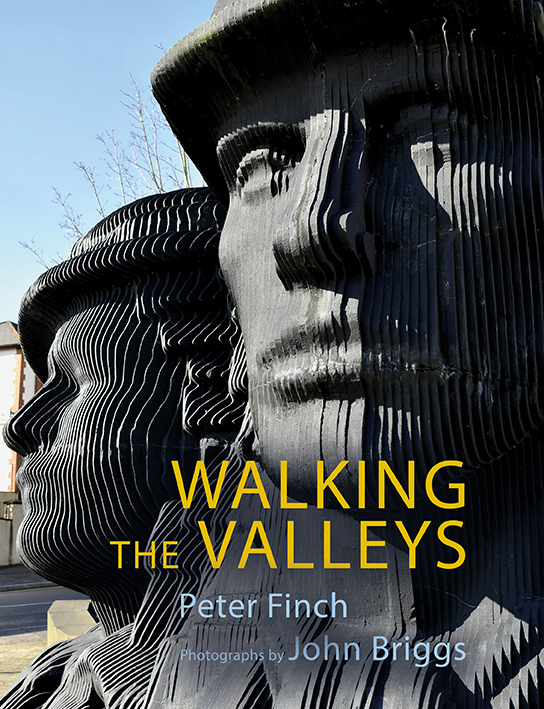 This cover shows a photo of a large sculpture depicting the heads of two miners made from horizontal black slabs. The text reads: Walking the Valleys. Peter Finch. Photographs by John Briggs.