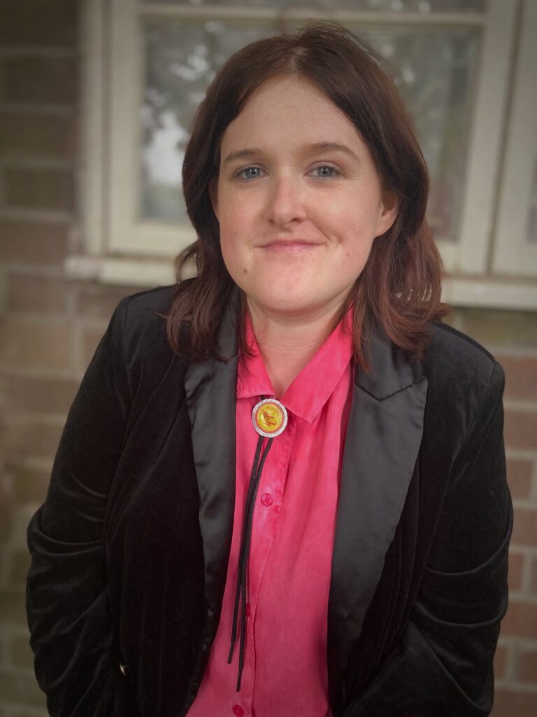 Photo of Rhian Elizabeth smiling at the camera. She has shoulder length brown hair and is wearing a pink shirt and black blazer.