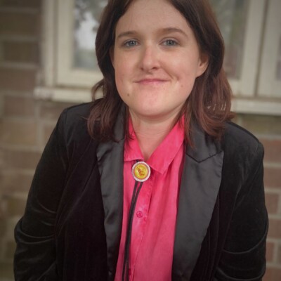 Photo of Rhian Elizabeth smiling at the camera. She has shoulder length brown hair and is wearing a pink shirt and black blazer.
