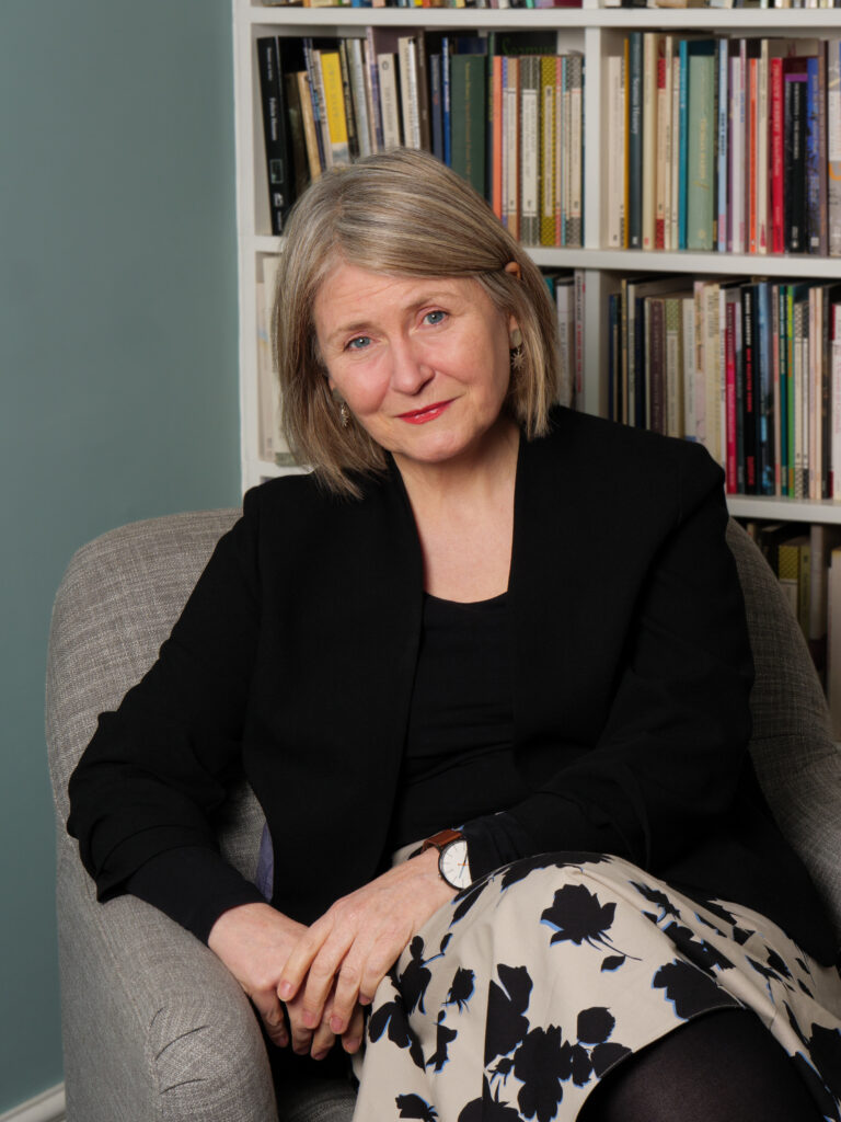 Photo of Deryn Rees-Jones sat in a chair in front of a neat bookshelf. She is wearing a black jacket and printed skirt.