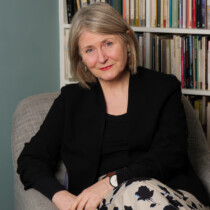 Photo of Deryn Rees-Jones sat in a chair in front of a neat bookshelf. She is wearing a black jacket and printed skirt.