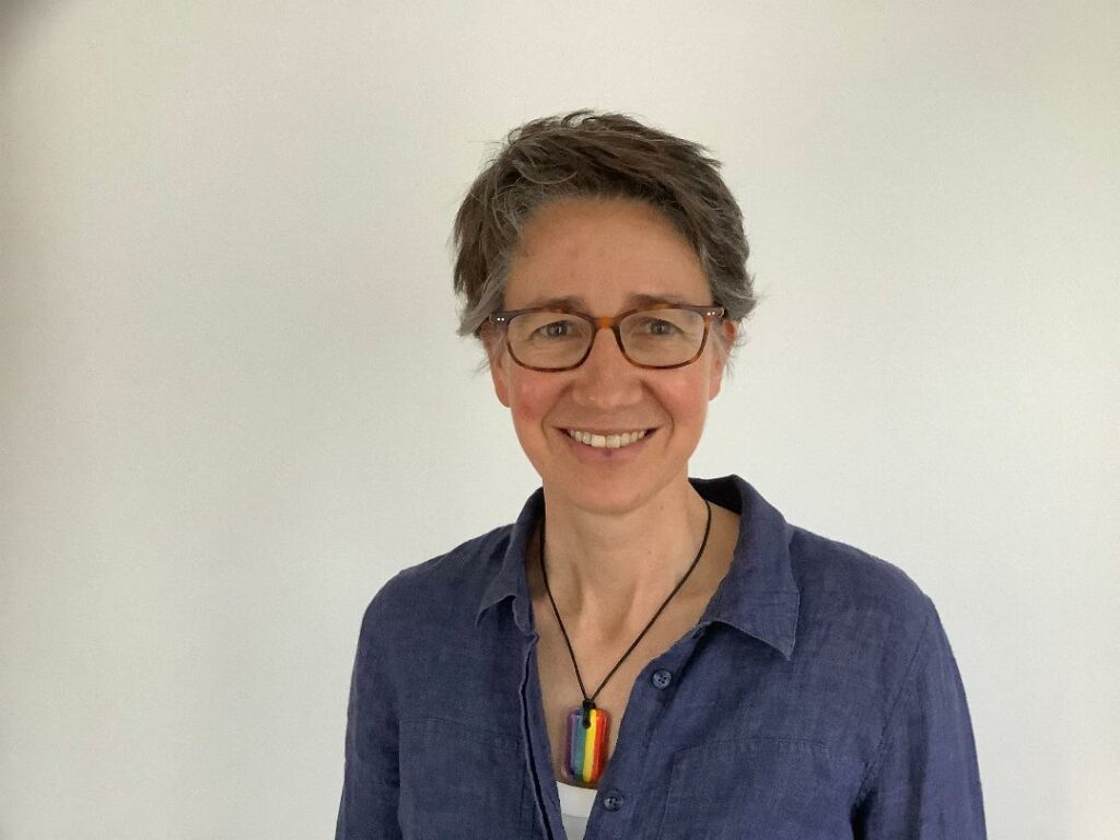 Photo of Alison Binney standing in front of a white wall. She is smiling and is wearing a blue shirt and glasses.