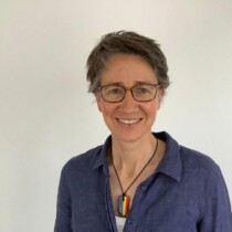 Photo of Alison Binney standing in front of a white wall. She is smiling and is wearing a blue shirt and glasses.