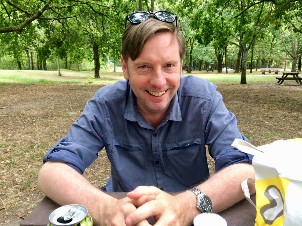 Photograph of Andrew Taylor sitting at a picnic table in a park. He is wearing a blue shirt and is smiling at the camera.