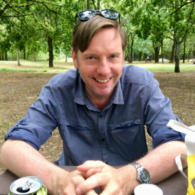 Photograph of Andrew Taylor sitting at a picnic table in a park. He is wearing a blue shirt and is smiling at the camera.