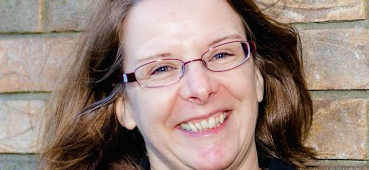 Anne Lauppe-Dunbar is a white woman with short-medium light brown hair. She is wearing glasses with clear lenses and a dark blue shirt. She is looking into the camera and smiling.