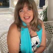 Photo of Maggie Harris wearing a blue scarf and a black and white floral top. She has shoulder length hair and is smiling at the camera.