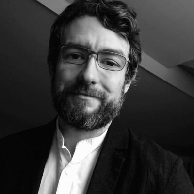 Black and white photo of Ben Wilkinson, a white man with short hair and a beard. He is wearing glasses, a light shirt and jacket