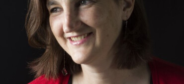 Cath Drake is a white woman with short dark hair. She is wearing a black top and red cardigan.