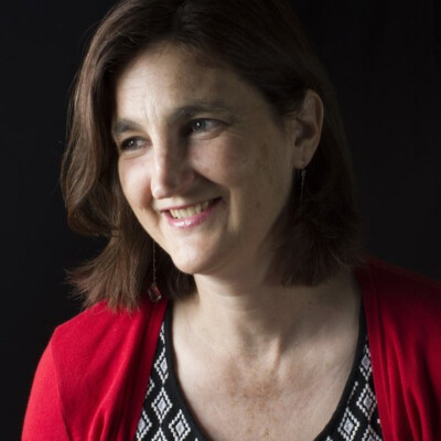 Cath Drake is a white woman with short dark hair. She is turned slightly to the right of the camera and is smiling. She is wearing a red cardigan with a black top underneath.