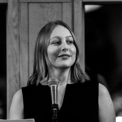 Black and white photo of Elizabeth Parker. She is a white woman with shoulder length hair. She is standing in front of a microphone, looking to the right of the camera and smiling.