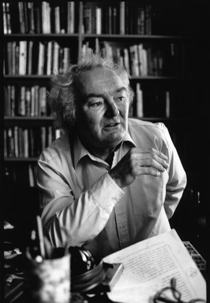 Black and white photo of John Ormond. He is a white man with light curly hair, wearing a shirt. He is looking toward the right of the camera with his right arm in front of his body. In the background are books of shelves.