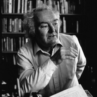 Black and white photo of John Ormond. He is a white man with light curly hair, wearing a shirt. He is looking toward the right of the camera with his right arm in front of his body. In the background are books of shelves.
