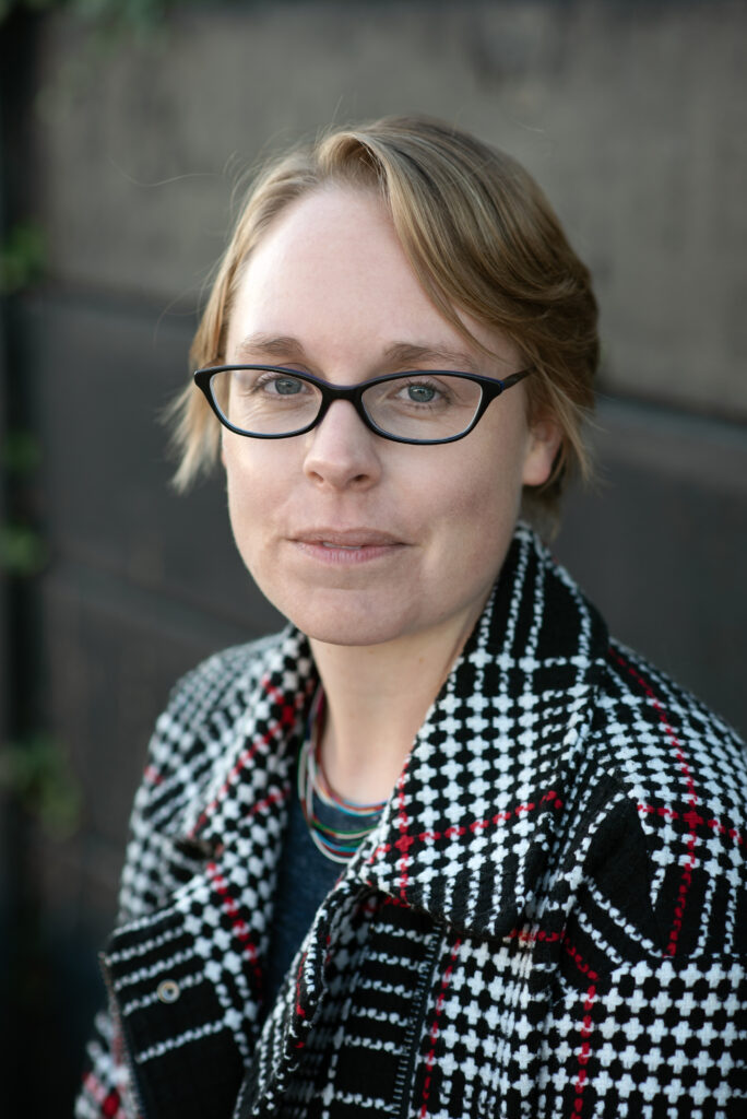 Katherine Stansfield is white woman with short blonde hair which stops past her ears. She is wearing glasses and looking at the camera. She is wearing a dark coat with a white pattern on the collar.