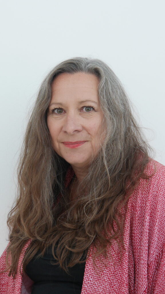 Kate Noakes is a white woman with long hair. She is wearing a black top and pink cardigan.