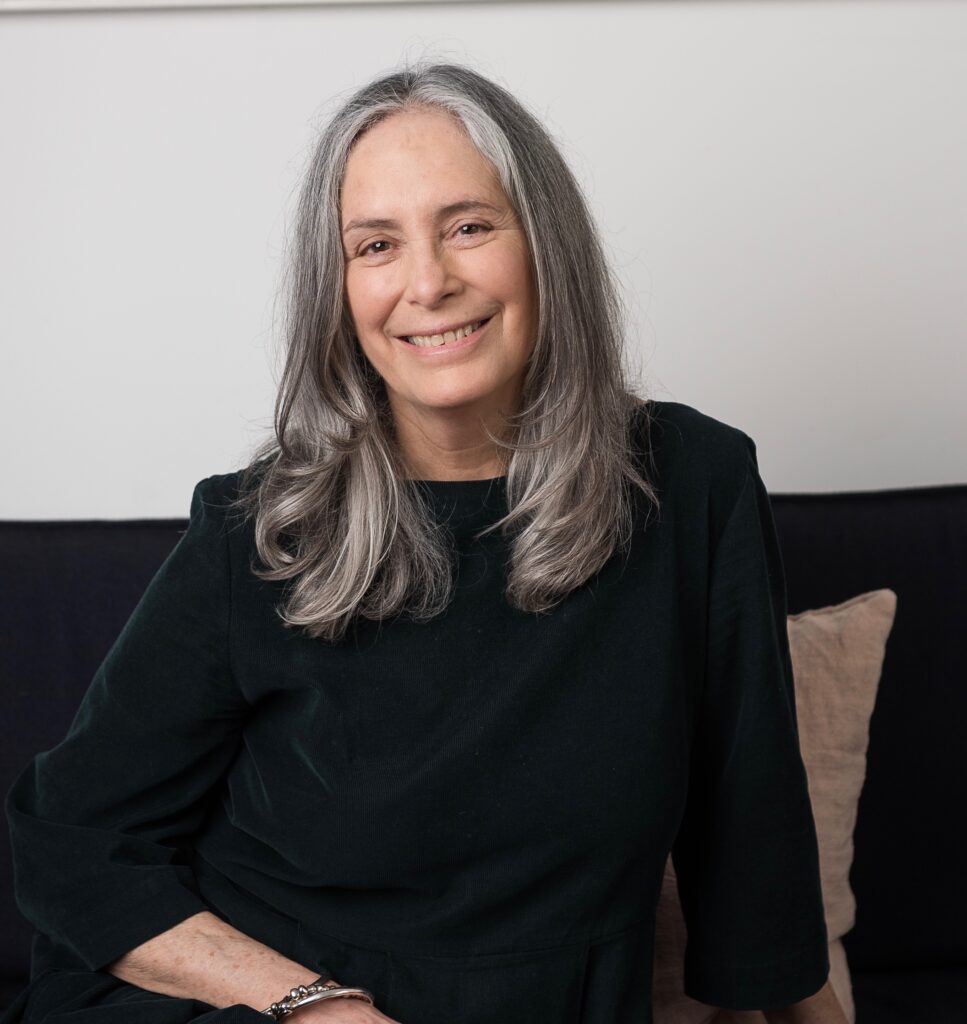 Lynne Hjemlgaard is a white woman with light hair that stops past her shoulders. She is looking into the camera and smiling. She is wearing a black top and sitting on a dark sofa.
