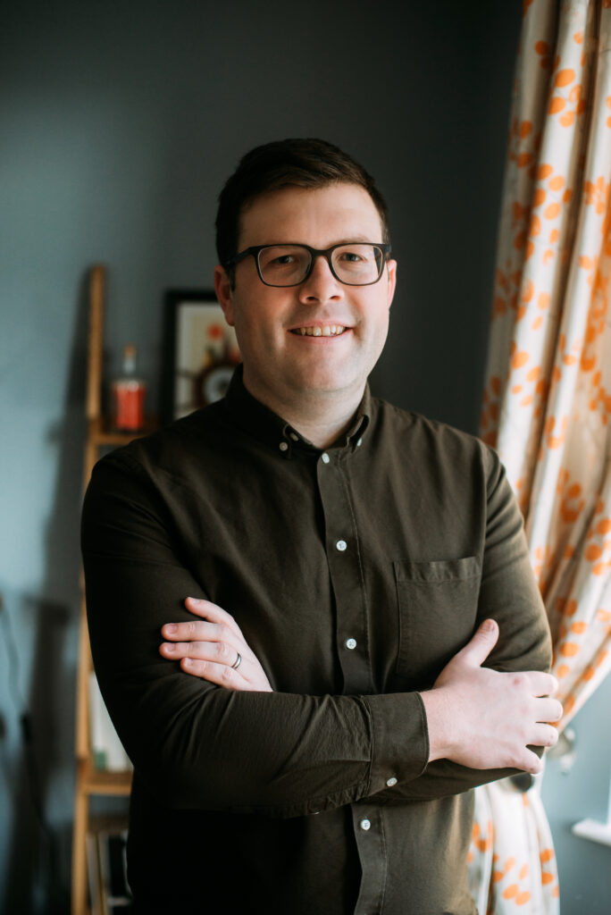 Nathan Munday is a white man with short dark hair. He is standing with his arms crossed loosely in front of him and is smiling at the camera. He is wearing glasses and a green shirt