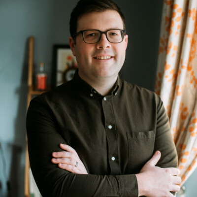 Nathan Munday is a white man with short dark hair. He is standing with his arms crossed loosely in front of him and is smiling at the camera. He is wearing glasses and a green shirt