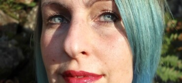 Polly Atkin is a white woman with short hair dyed blue. She is standing outside in the sun and is looking at the camera.