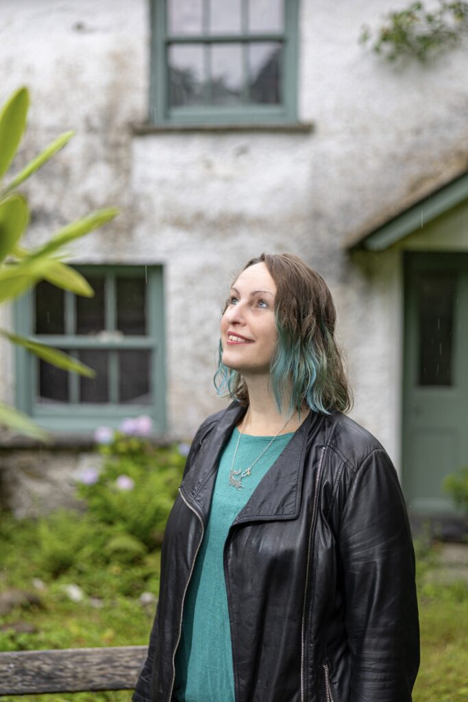 Photo of Polly Atkin standing outside and looking up at the sky. She is wearing a black jacket and turquoise top.