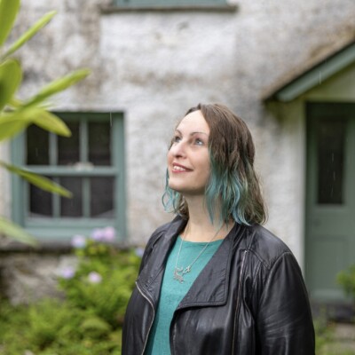Photo of Polly Atkin standing outside and looking up at the sky. She is wearing a black jacket and turquoise top.