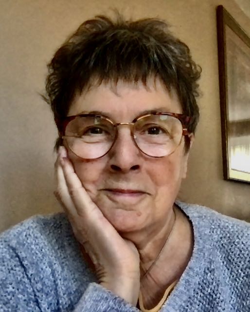 Rachael Clyne is a white woman with short dark hair. She is leaning her cheek against one hand and is smiling at the camera. She is wearing glasses and a light grey jumper.