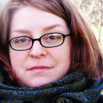 Rhiannon Hooson is a white woman with shoulder-length red hair. She is wearing glasses and a blue scarf.
