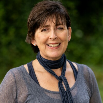 Sarah Wimbush is a white woman with short dark hair. She has a thin blue scarf around her neck, a dark blue top and a slightly translucent shirt over her dark blue top. She is looking into the camera and smiling.