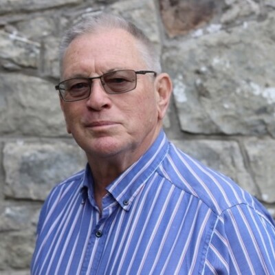 Tony Curtis is a white man with short light hair and glasses. He is wearing a blue striped shirt.