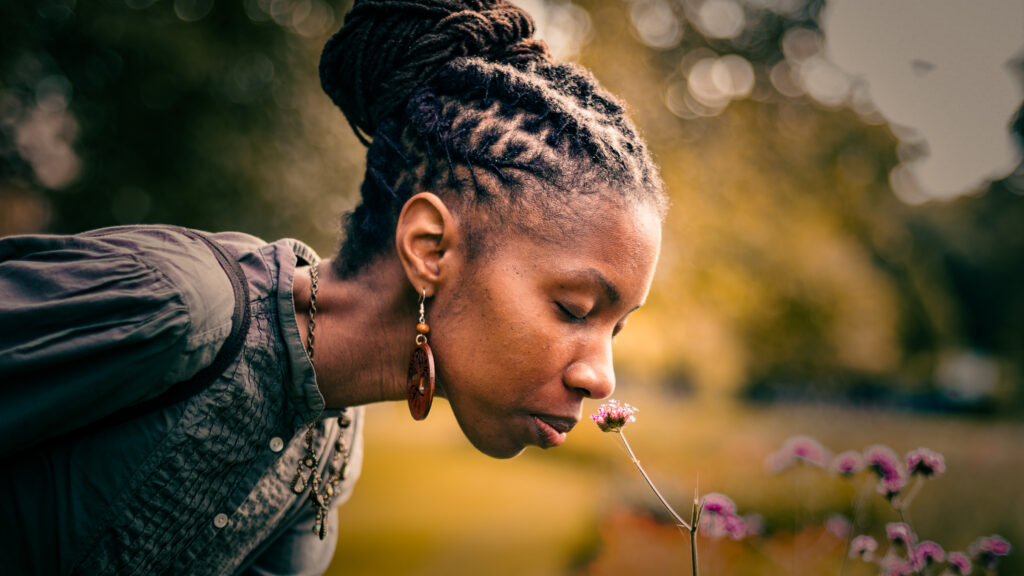 Photo of Zakia Carpenter-Hall bending down to smell a small pink flower. Her hair is braided and tied back into a bun.