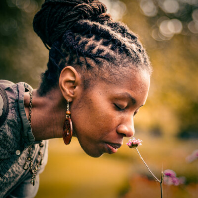 Photo of Zakia Carpenter-Hall bending down to smell a small pink flower. Her hair is braided and tied back into a bun.