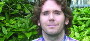 Jonathan Edwards has dark curly hair and is wearing a checked purple shirt. He is standing outside and is smiling at the camera.