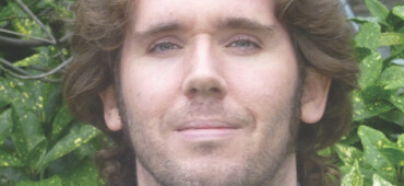 Jonathan Edwards has dark curly hair and is wearing a checked purple shirt. He is standing outside and is smiling at the camera.