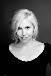Black and white photo of Kathryn Gray. She has light skin and white hair which contrasts her dark top. She is looking into the camera and smiling.