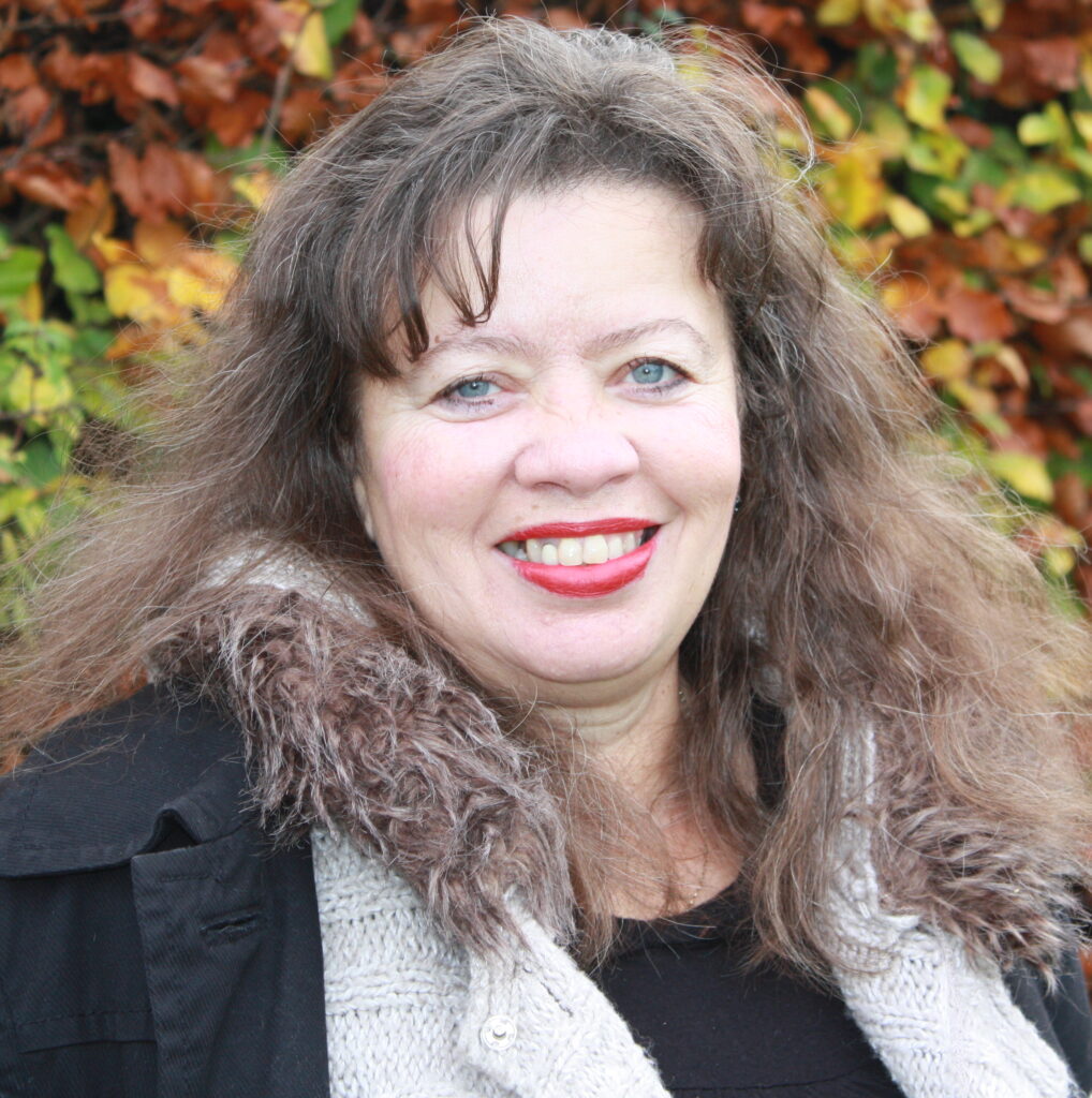 Photo of Maggie Harris. Maggie has slightly curly brown hair that is just past shoulder length. She is wearing red lipstick and black coat with a fur trim. She is looking at the camera and smiling.