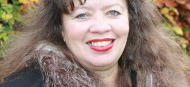 Photo of Maggie Harris. Maggie has slightly curly brown hair that is just past shoulder length. She is wearing red lipstick and black coat with a fur trim. She is looking at the camera and smiling.