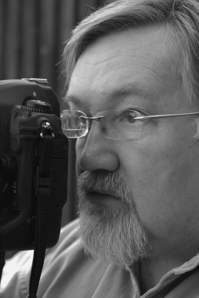 Close up black and white photo of Phil Cope. He is holding a camera to his eye and looking through the lens.