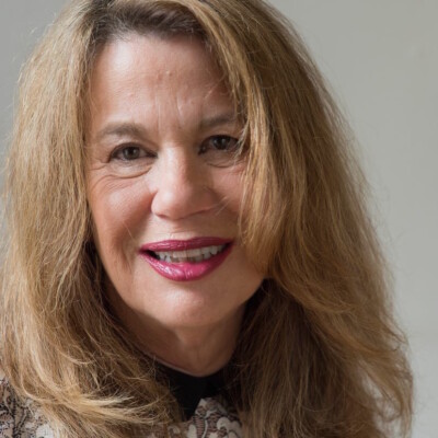 Sue Hubbard is a white woman with shoulder-length blonde hair. She is wearing red lipstick and a dark top. She is looking into the camera and smiling.