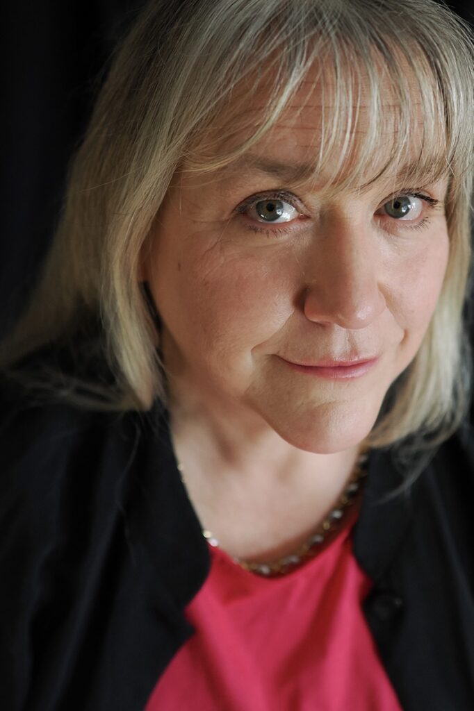 Close up photo of Tiffany Murray looking up at the camera. She has shoulder-length blonde hair and a fringe and is wearing a pink top and black cardigan.