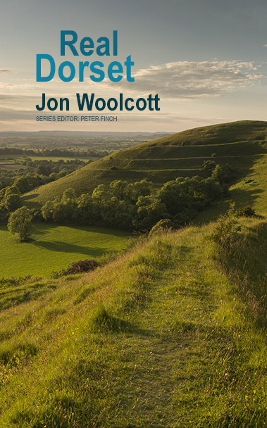 This cover shows a photo of a Dorset Hill Fort. A lush grass path leads to the domed fort in the distance which sits beneath a blue sky tinged with the golden light of early dusk. The text reads: Real Dorset. Jon Woolcott.