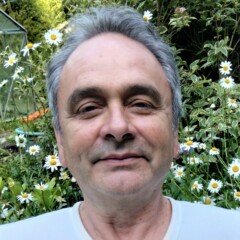 Photo of Simon Hicks standing in a garden and smiling. He is wearing a white t-shirt.
