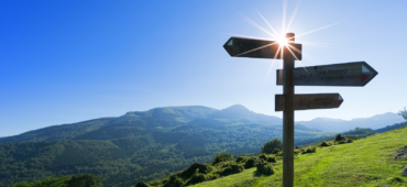 Photo of a wooden sign post pointing in various directions. Blue sky and green hills roll away behind it.