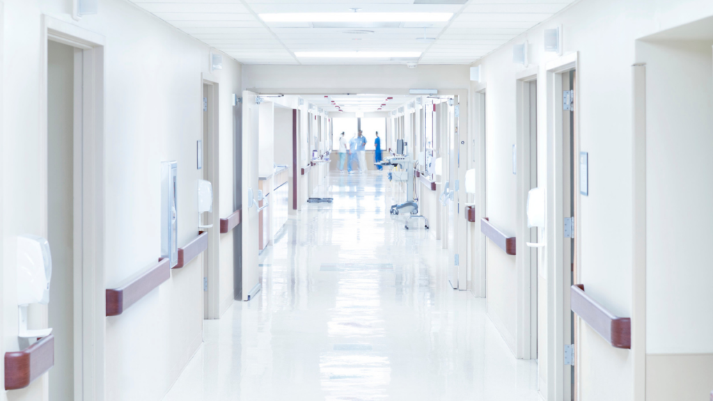 An empty hospital corridor. At the far end are some blurry people dressed in blue scrubs.