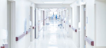 An empty hospital corridor. At the far end are some blurry people dressed in blue scrubs.