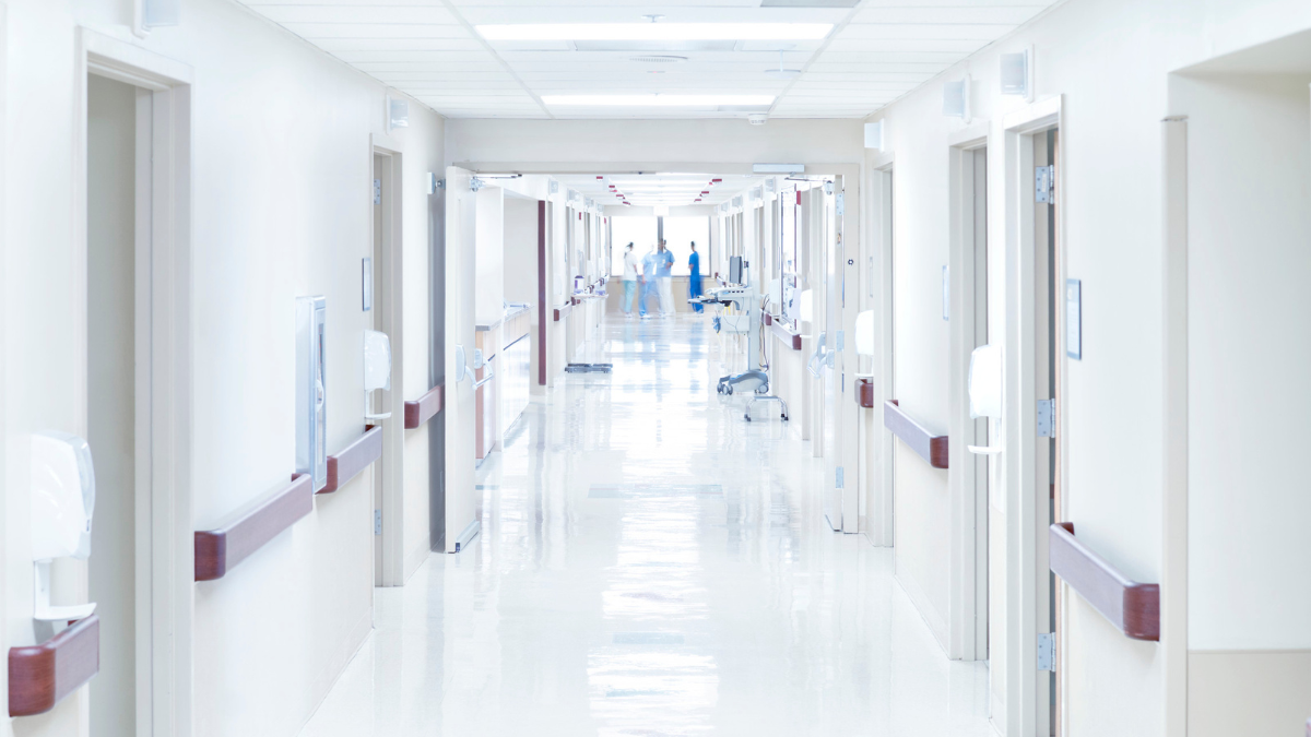An empty hospital corridor. At the far end are some blurry people dressed in blue scrubs.
