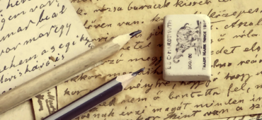 Photo of papers covered in cursive handwriting laying in a pile with two pencils and a rubber on top