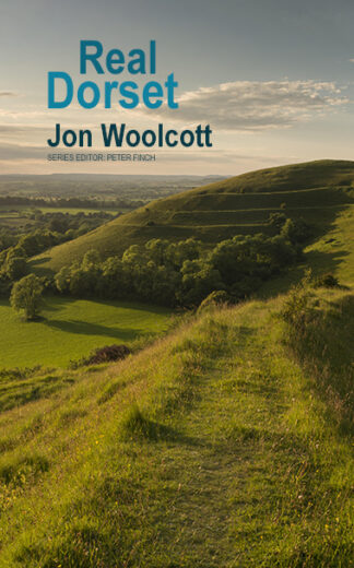 This cover shows a photo of a Dorset Hill Fort. A lush grass path leads to the domed fort in the distance which sits beneath a blue sky tinged with the golden light of early dusk. The text reads: Real Dorset. Jon Woolcott.