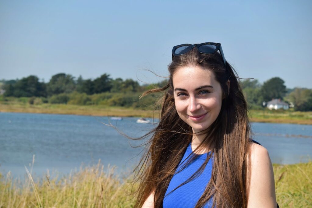 Photo of Bethany Handley outside by an open lake in the sunshine. She has long dark hair held back with sunglasses and is wearing a blue top.