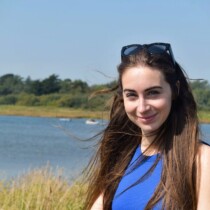 Photo of Bethany Handley outside by an open lake in the sunshine. She has long dark hair held back with sunglasses and is wearing a blue top.