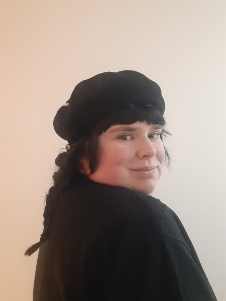 Betty Doyle is a white woman with long dark hair. She is wearing a black top and beret and is looking over her shoulder towards the camera.
